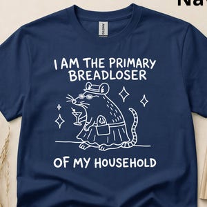 May include: A navy blue short-sleeved t-shirt featuring a white line-art graphic of a rat wearing a hat and sunglasses, holding a martini glass and a loaf of bread. The text reads: I AM THE PRIMARY BREADLOSER OF MY HOUSEHOLD.