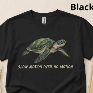 Puede incluir: Una camiseta negra de manga corta con una ilustración realista de una tortuga marina verde nadando. Debajo de la tortuga, el texto SLOW MOTION OVER NO MOTION aparece escrito en una fuente sans-serif de color tostado.