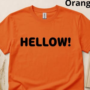 May include: A bright orange short-sleeved t-shirt featuring the word HELLOW! printed in bold black capital letters across the centre of the chest. The shirt has a crew neckline and a standard fit, with a small white brand label at the inner neck.