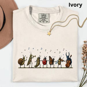 May include: An ivory t-shirt featuring a graphic of various insects playing musical instruments, including a snail, grasshopper, caterpillar, ladybug, butterfly, bee, and beetle. The shirt has a Comfort Colors label and a gold star necklace.