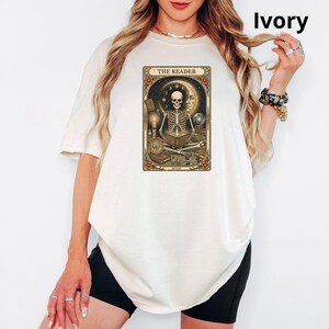 Op de afbeelding: Ivoor kleurig T-shirt met een tarotkaart ontwerp. De kaart toont een skelet dat een boek leest, met de tekst "THE READER". Het ontwerp bevat hemelse en mystieke elementen. Het T-shirt is een casual stijl.