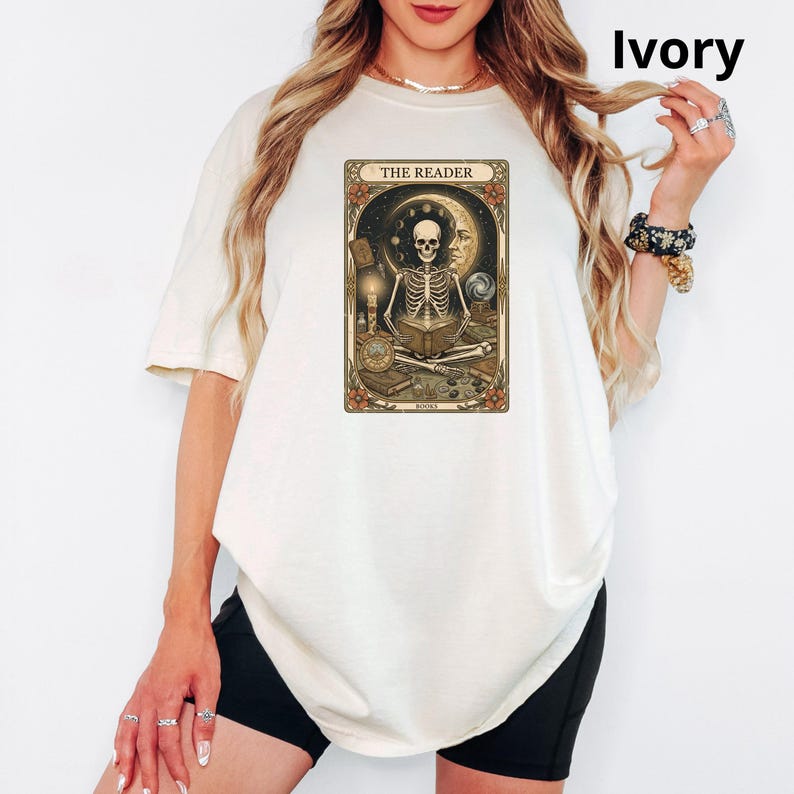 Op de afbeelding: Ivoor kleurig T-shirt met een tarotkaart ontwerp. De kaart toont een skelet dat een boek leest, met de tekst "THE READER". Het ontwerp bevat hemelse en mystieke elementen. Het T-shirt is een casual stijl.