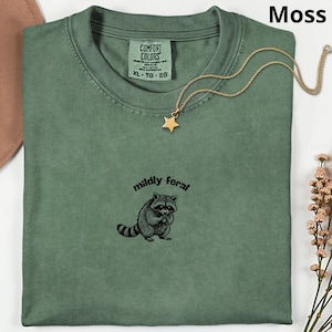 Pode incluir: Camiseta verde musgo com gola redonda e mangas curtas. A camiseta apresenta um gráfico de guaxinim em preto e branco com as palavras "mildly feral". Um colar com pingente de estrela dourada repousa sobre a camiseta.