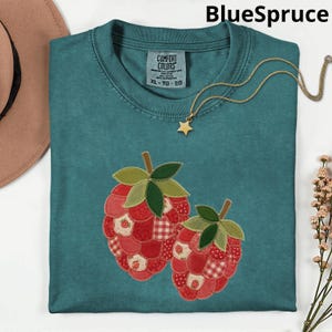 Peut inclure: T-shirt XL Comfort Colors bleu sarcelle avec un motif cousu de deux framboises rouges et de feuilles vertes. Un collier doré avec un pendentif étoile et un chapeau marron sont également visibles. Le texte "BlueSpruce" est en haut.