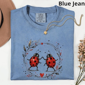 May include: A pale blue t-shirt featuring a design of two red ladybirds holding hands within a wreath. A gold star pendant necklace is draped on the shirt. The shirt is labelled "Comfort Colors" and is size XL-TG-EG.