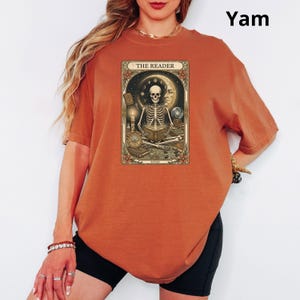 Op de afbeelding: Verbrand oranje t-shirt met een tarotkaartontwerp. De kaart toont een skelet dat een boek leest, met de tekst "THE READER" bovenaan. Een comfortabele, casual stijl.