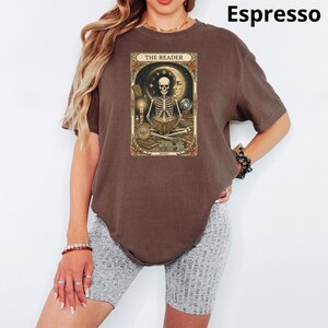 Op de afbeelding: Espresso-kleurig t-shirt met een tarotkaartontwerp met een skelet dat een boek leest, de tekst "THE READER" staat bovenaan. Het shirt is een comfortabele, casual stijl.