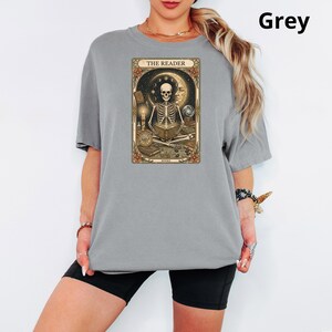 Op de afbeelding: Grijze t-shirt met een tarotkaart ontwerp. De kaart toont een skelet dat een boek leest, tegen een achtergrond van maan en sterren. De tekst "THE READER" staat bovenaan de kaart. Het woord "BOOKS" staat onderaan de kaart.
