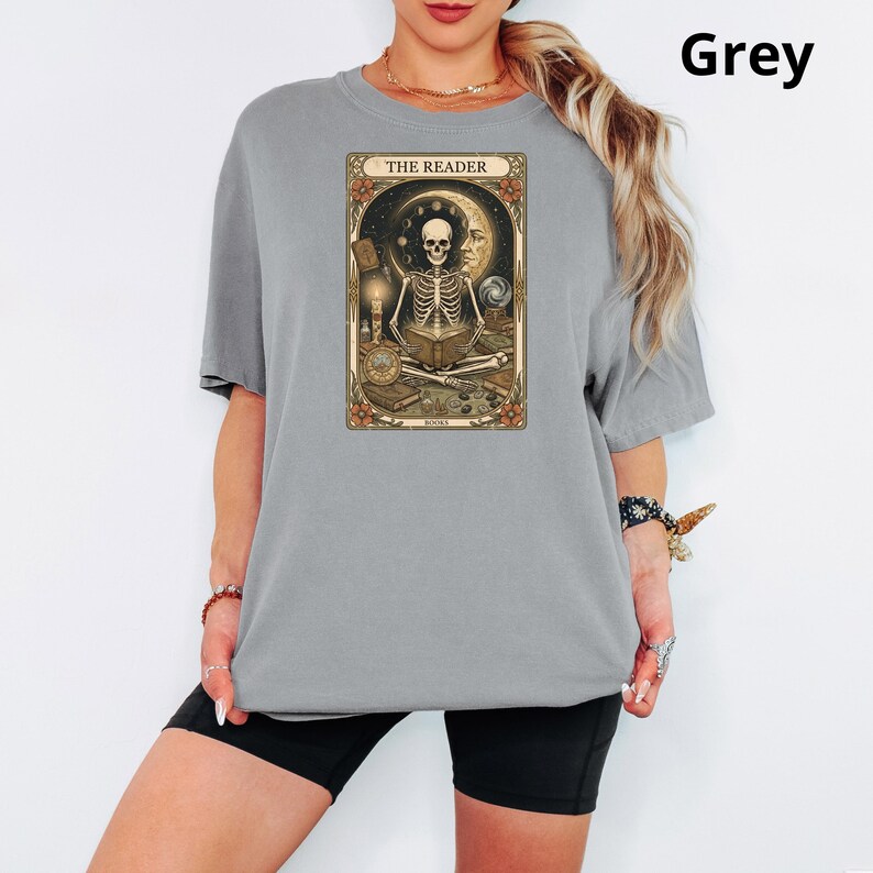 Op de afbeelding: Grijze t-shirt met een tarotkaart ontwerp. De kaart toont een skelet dat een boek leest, tegen een achtergrond van maan en sterren. De tekst "THE READER" staat bovenaan de kaart. Het woord "BOOKS" staat onderaan de kaart.