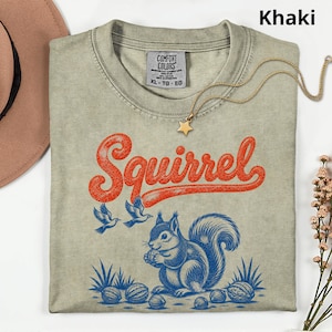May include: A khaki-colored t-shirt with a Comfort Colors label at the neck. The graphic features the word Squirrel in orange cursive script above a blue illustration of a squirrel holding a nut, surrounded by two birds and several walnuts.