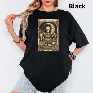 Op de afbeelding: Zwart T-shirt met een tarotkaartontwerp. De afbeelding toont een skelet dat een boek leest, met de tekst "THE READER" bovenaan. Het ontwerp bevat een maan, sterren en bloemmotieven. De algehele esthetiek is gotisch en mystiek.