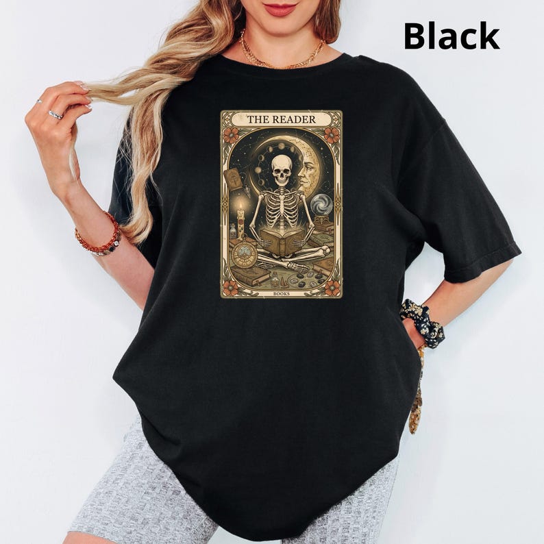 Op de afbeelding: Zwart T-shirt met een tarotkaartontwerp. De afbeelding toont een skelet dat een boek leest, met de tekst "THE READER" bovenaan. Het ontwerp bevat een maan, sterren en bloemmotieven. De algehele esthetiek is gotisch en mystiek.