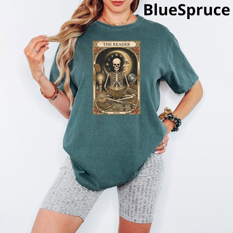 Op de afbeelding: Tealkleurig T-shirt met een tarotkaartontwerp met een skelet dat een boek leest, getiteld "THE READER". De afbeelding bevat een halve maan, sterren en diverse objecten. Het shirt heeft een comfortabele, oversized pasvorm.