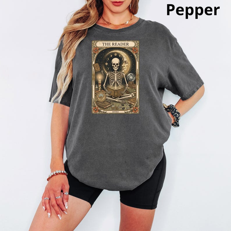 Op de afbeelding: Donkergrijs T-shirt met een tarotkaartontwerp met een skelet dat een boek leest, een maan en de tekst "THE READER". Het shirt heeft korte mouwen en een relaxte pasvorm.