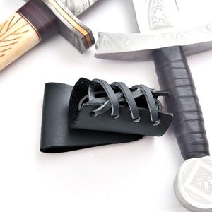 May include: Close-up of a black leather sword scabbard with decorative stitching. Two swords are visible in the background, one with a bone handle and the other with a silver blade and black grip. The swords are on a white background.