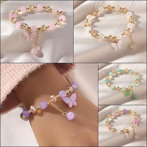 Könnte beinhalten: Eine Sammlung von Armbändern mit Perlen und Schmetterlingsanhängern. Die Armbänder weisen verschiedene Farben von Perlen auf, darunter Rosa, Weiß, Lila und Grün, mit goldfarbenen Akzenten. Jedes Armband hat einen hängenden Schmetterlingsanhänger.