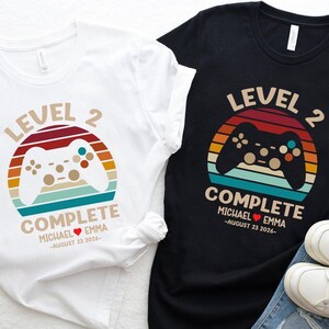 May include: Two t-shirts, one white and one black, with a video game controller design. The design includes the words "Level 2 Complete" and a couple's names and wedding date. The shirts are paired with blue jeans and white sneakers.