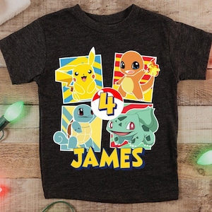 May include: Dark gray t-shirt featuring four colorful cartoon characters, the number 4, and the name "JAMES" in yellow. The shirt has a crew neck and short sleeves. Christmas lights and a Santa hat are in the background.