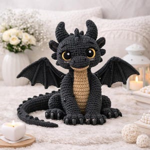 以下が含まれることがあります： 手作りの編み物ドラゴン。濃いグレーで、お腹はベージュ、大きな黄色い目があります。ドラゴンは黒い翼、角、爪を持っています。白いふわふわした表面に座っています。気まぐれな装飾品です。