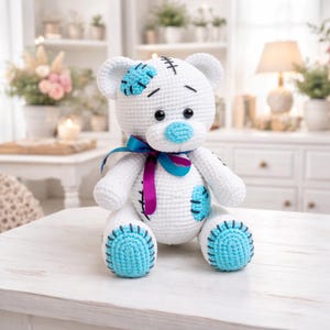 Puede incluir: Un oso de peluche blanco tejido a mano con detalles azules en las orejas, la nariz y los parches. El oso tiene detalles cosidos en negro y una cinta azul y morada alrededor del cuello. Está sentado sobre una superficie blanca.