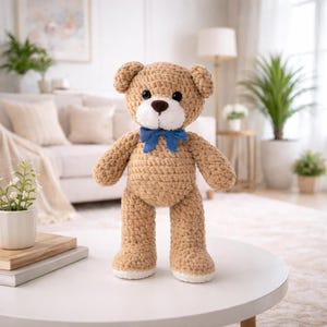 Puede incluir: Un oso de peluche de ganchillo marrón hecho a mano con una pajarita azul. El oso tiene ojos negros, hocico blanco y suelas blancas. El oso está de pie sobre una mesa blanca.