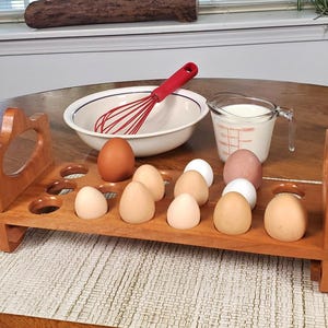 May include: A rectangular wooden egg holder with twelve circular cutouts, containing eleven eggs of varying shades of brown and white. Behind the holder sits a white mixing bowl with a red wire whisk and a glass measuring cup filled with milk.