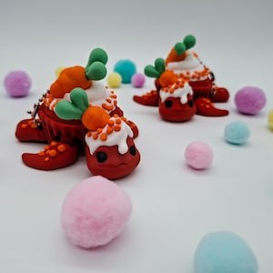 Peut inclure: Deux figurines rouges en forme de tortue avec des coquilles en forme de cupcake, surmontées de carottes oranges et de feuilles vertes. Les figurines sont entourées de pompons colorés dans les tons de rose, bleu, violet et jaune. Le fond est blanc.