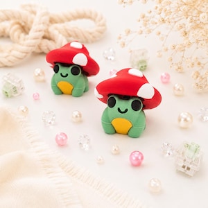 Può includere: Due piccole statuette di rane verdi con pancia gialla e occhi neri, ognuna con un cappello a forma di fungo rosso con macchie bianche. Le rane sono circondate da perle bianche e rosa, perline sfaccettate trasparenti e switch per tastiere meccaniche.