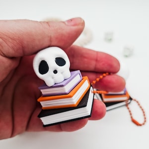 Könnte beinhalten: Ein weißer Totenkopf sitzt auf einem Stapel Miniaturbücher in Lila, Orange und Schwarz. Die Bücher haben weiße Seiten und sind an einer orangefarbenen Perlenkette befestigt. Das Thema ist Halloween oder Gothic.