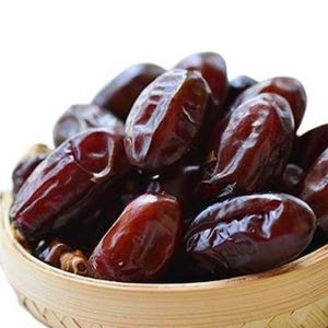 Puede incluir: Primer plano de una cesta tejida llena de dátiles de color marrón oscuro. Los dátiles tienen una textura brillante y arrugada y están apilados. La cesta es de madera de color claro.