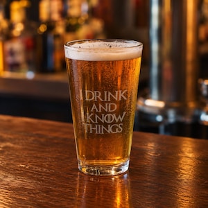 Könnte beinhalten: Ein klares Bierglas gefüllt mit einer goldgelben Flüssigkeit und einer weißen Schaumkrone. Das Glas zeigt den geätzten weißen Schriftzug I DRINK AND I KNOW THINGS mit einem kreisförmigen Symbol im Buchstaben O, zentriert auf der Vorderseite.