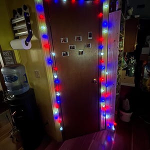 May include: A wooden door decorated with a string of glowing red, white, and blue spherical lights draped around the frame. Several small, rectangular white-bordered photographs are attached to the center of the brown door surface.