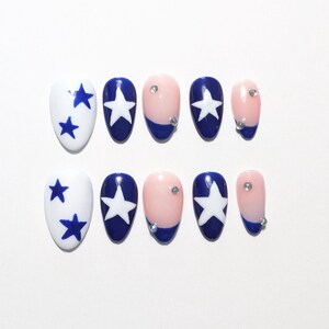 Peut inclure: Un ensemble de dix faux ongles au motif patriotique. Les designs incluent des ongles blancs avec des étoiles bleues, des ongles bleus avec des étoiles blanches, et des ongles nude avec des pointes françaises bleues ornées de strass.