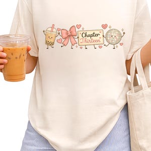 May include: A cream-colored t-shirt featuring a graphic print of a boba tea cup, a pink bow, a rectangular sign with the text "Chapter Thirteen," and a disco ball, all decorated with small pink hearts and sparkles on the front of the shirt.