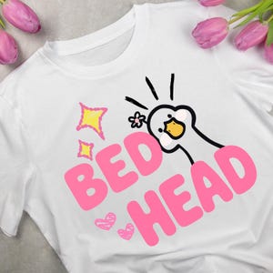 May include: A white t-shirt featuring a graphic of a cartoon goose with a small flower on its head. The text "BED HEAD" is printed in large pink letters. The design includes yellow star shapes and pink hand-drawn hearts surrounding the goose.