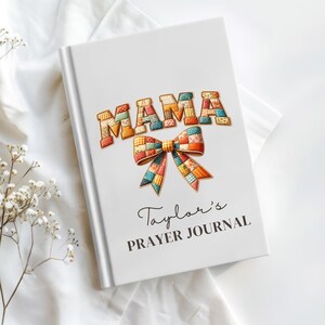 May include: White journal with the word "MAMA" in patchwork letters and a patchwork bow illustration. The text "Taylor's PRAYER JOURNAL" is below the bow. The journal is on a white surface.