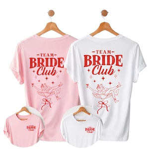 Può includere: T-shirt rosa e bianche con la scritta "TEAM BRIDE CLUB" in rosso. Le magliette presentano una grafica di una donna in un bicchiere da cocktail. Sono visibili anche magliette più piccole abbinate.