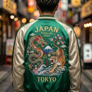 Puede incluir: Una chaqueta bomber de satén verde con mangas color crema y cuello acanalado. La parte posterior presenta un bordado detallado de un dragón, un tigre, el monte Fuji, flores de cerezo y olas, con el texto JAPAN SUKAJAN 2024 y TOKYO.