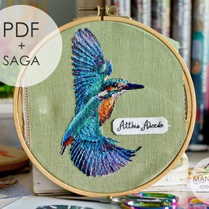 May include: A cross-stitch embroidery of a kingfisher bird in flight, featuring blue, teal, and orange hues. The artwork is set within a wooden embroidery hoop against a light green fabric. The text "PDF + SAGA" is in a white circle.