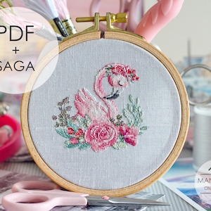 May include: A circular wooden embroidery hoop containing light gray fabric with a cross-stitch design of a pink flamingo surrounded by pink and red flowers and green leaves. The text PDF + SAGA is visible on the left, and MANSARDA cross stitch is on the right.