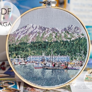 May include: A circular wooden embroidery hoop containing a cross-stitch pattern of a harbor with fishing boats, a dark green forest, and purple-toned mountains. The text PDF + SAGA is visible in the upper left, and MANSARDA cross stitch is at the bottom.