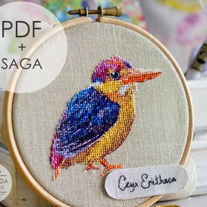 May include: A colorful cross-stitch embroidery of a bird, set in a wooden hoop. The bird features blue, yellow, and purple hues, with the text "PDF + SAGA" and "Ceyx Erithaca" visible. The brand logo "MANSARDA cross stitch" is also present.