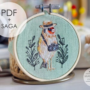 May include: A cross-stitch embroidery hoop featuring a bird wearing a hat and holding a camera. The bird is stitched in shades of brown, yellow, and white, set against a light green background. The text "PDF + SAGA" is in a white circle.