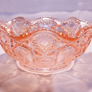 Op de afbeelding: Een roze, decoratieve glazen kom met een geschulpte rand. De kom heeft ingewikkelde patronen van sterren en geometrische vormen. De kom staat op een wit oppervlak.