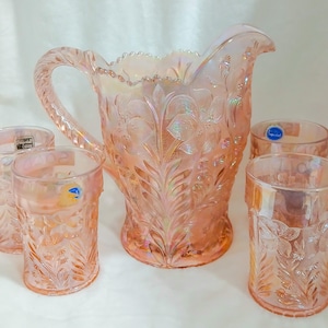 May include: A vintage pink iridescent glass pitcher and four matching glasses. The set features a floral pattern and a textured surface. The pitcher has a large handle and a spout. The glasses are cylindrical.