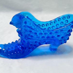 May include: A vibrant blue glass shoe figurine featuring a textured hobnail pattern across its surface. The decorative object is shaped like a high-heeled boot with a pointed toe and a raised opening, showcasing a translucent finish throughout.