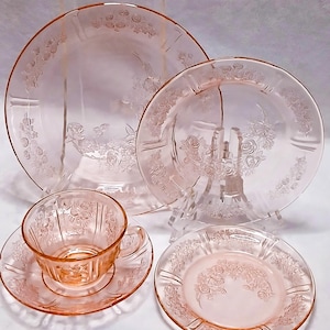 Puede incluir: Un conjunto de vajilla de vidrio rosa, que incluye platos, un platillo y una taza de té. Las piezas presentan un patrón floral de rosas en los bordes. El conjunto se exhibe sobre una superficie blanca.