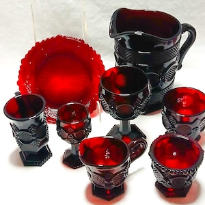 May include: A collection of ruby red glassware, including a pitcher, plate, and various glasses. The pieces have a textured pattern and are arranged on a white surface. The glassware is likely vintage.