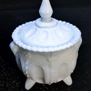 May include: A small, light blue milk glass jar with a matching lid. The jar features a raised beaded pattern, vertical arches, and three short legs. The lid has a pointed finial and a beaded edge, creating a textured, semi-translucent appearance.