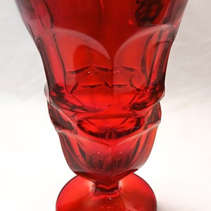 Puede incluir: Un jarrón de cristal rojo rubí con la parte superior acampanada y base redonda. El jarrón tiene un diseño detallado y texturizado alrededor del cuerpo. El cristal transparente permite el paso de la luz. Probablemente para fines decorativos.